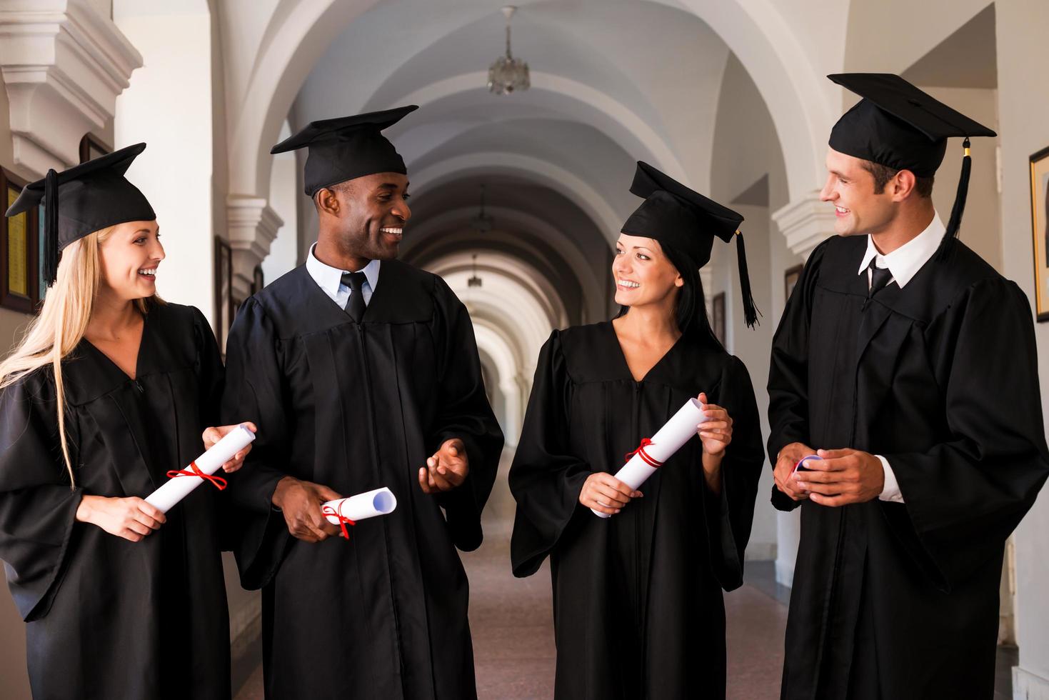 über eine glänzende Zukunft sprechen. Vier Hochschulabsolventen in Abschlusskleidern, die den Universitätskorridor entlanggehen und sich unterhalten foto