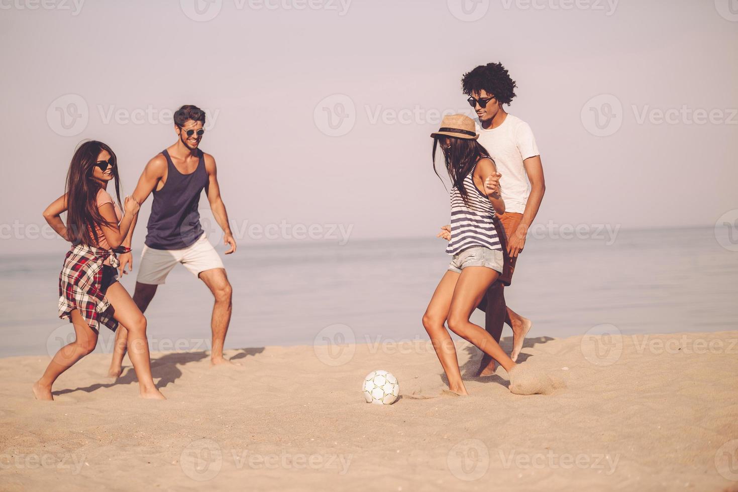 Strandspaß mit Freunden. gruppe fröhlicher junger leute, die mit fußball am strand mit meer im hintergrund spielen foto