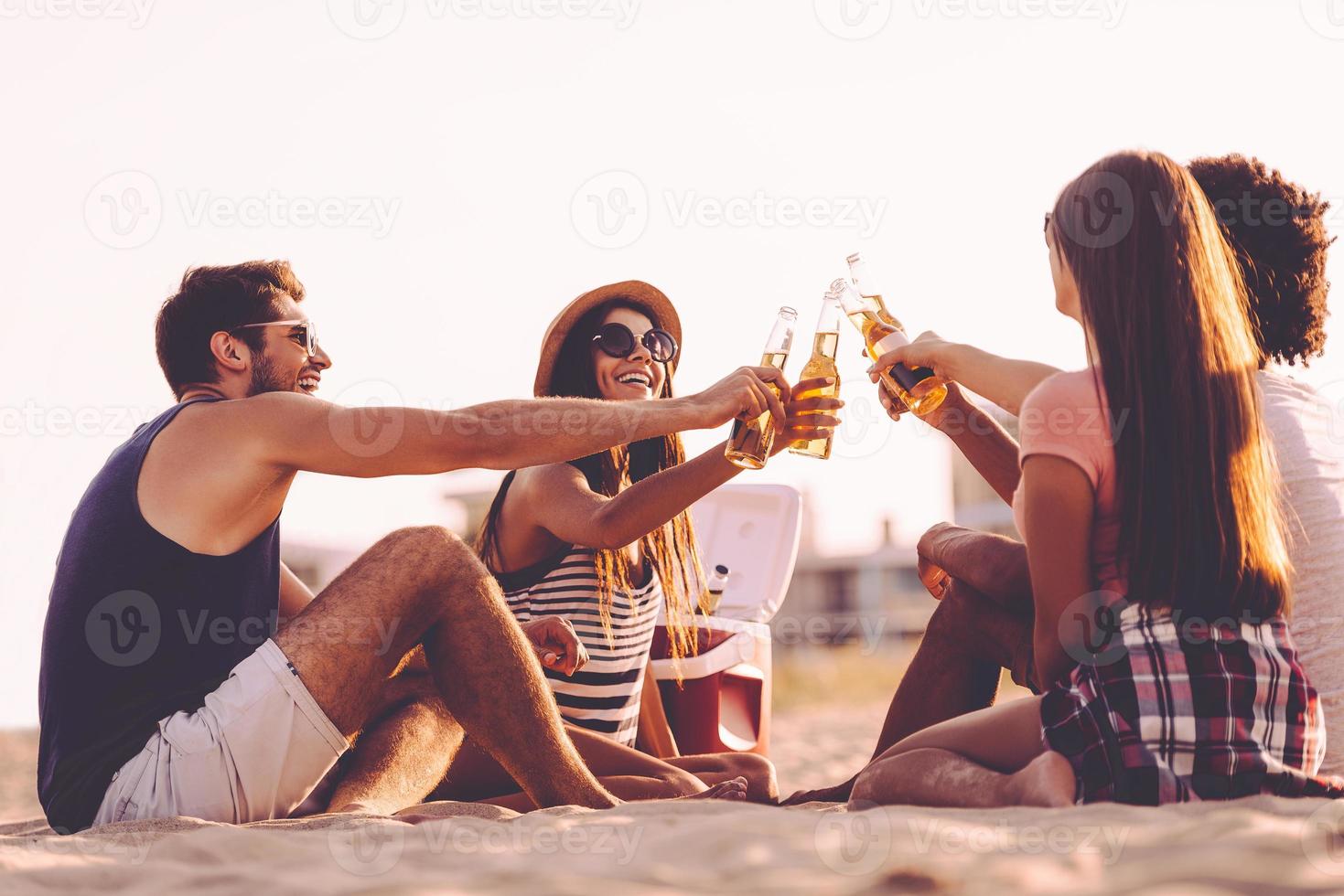 Prost fröhliche junge Leute, die mit Bierflaschen anstoßen, während sie ein Picknick am Strand genießen foto