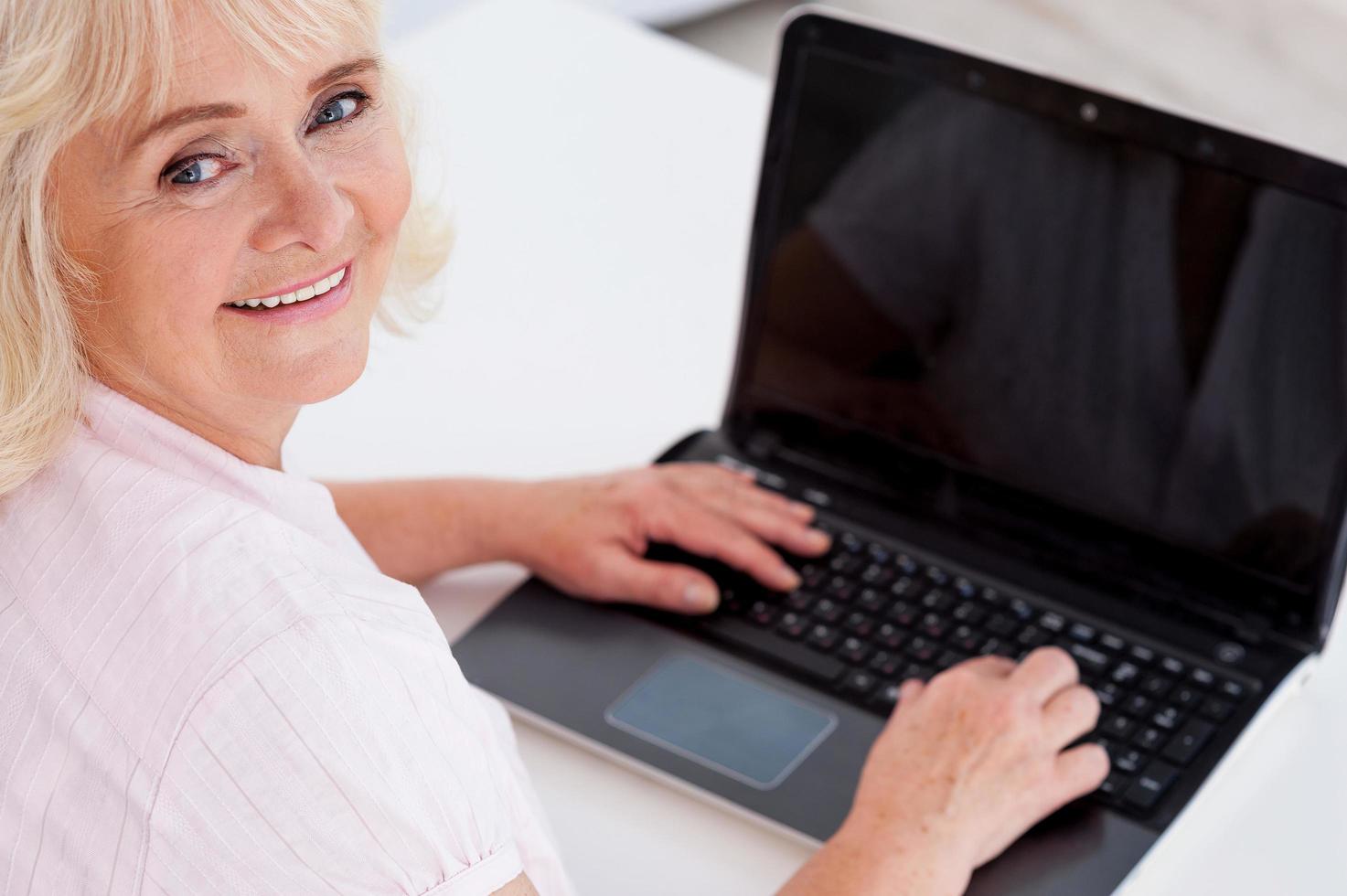 Senior im digitalen Zeitalter. Draufsicht einer fröhlichen Seniorin, die in die Kamera schaut und lächelt, während sie am Laptop arbeitet foto