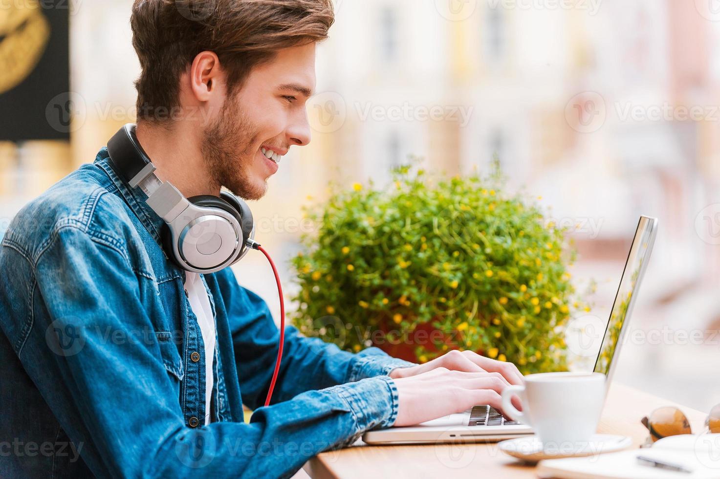 mit Freude arbeiten. Seitenansicht eines lächelnden jungen Mannes, der am Laptop arbeitet, während er im Straßencafé sitzt foto