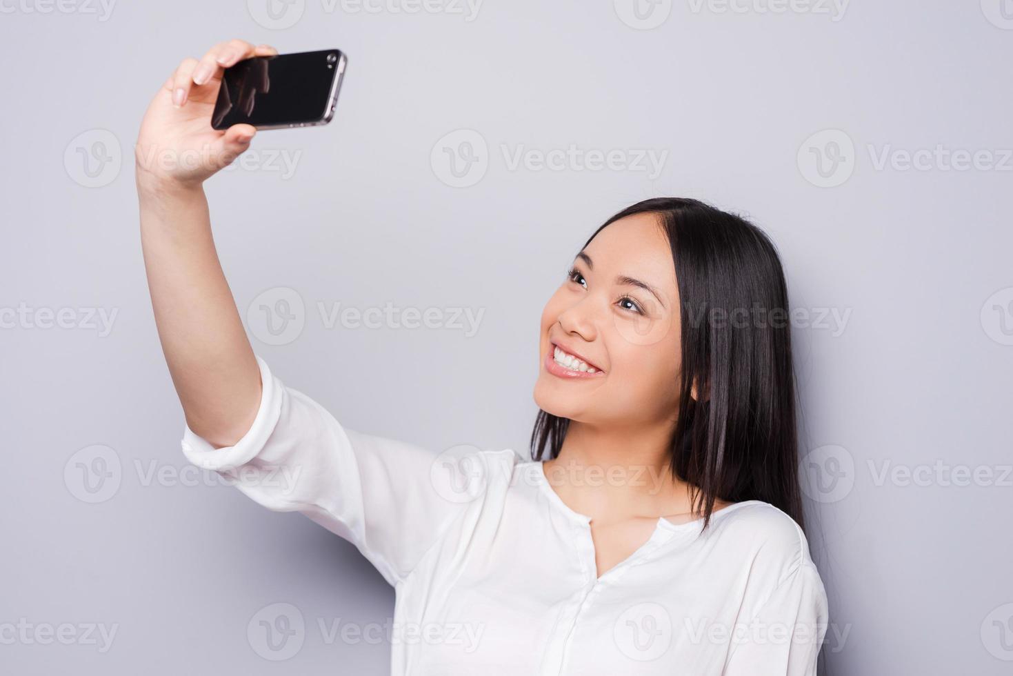 selfie fröhliche junge asiatische frau, die handy hält und selfie durch ihr intelligentes telefon macht, während sie gegen grauen hintergrund steht foto