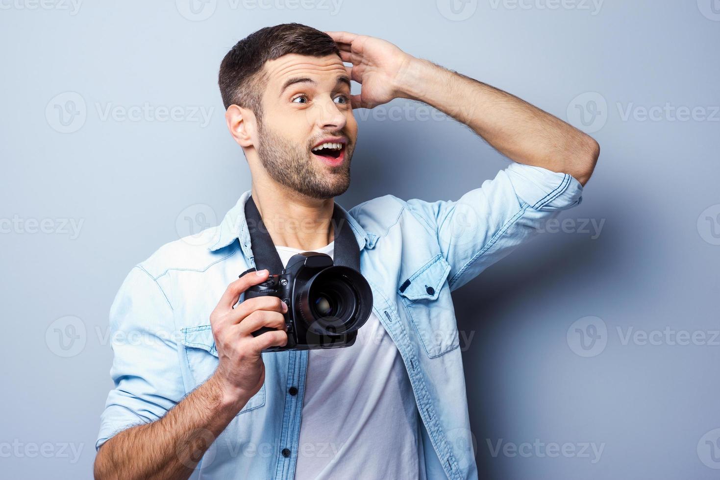 das ist einfach ein erstaunlich aufgeregter junger mann, der eine digitalkamera hält und wegschaut, während er vor grauem hintergrund steht foto