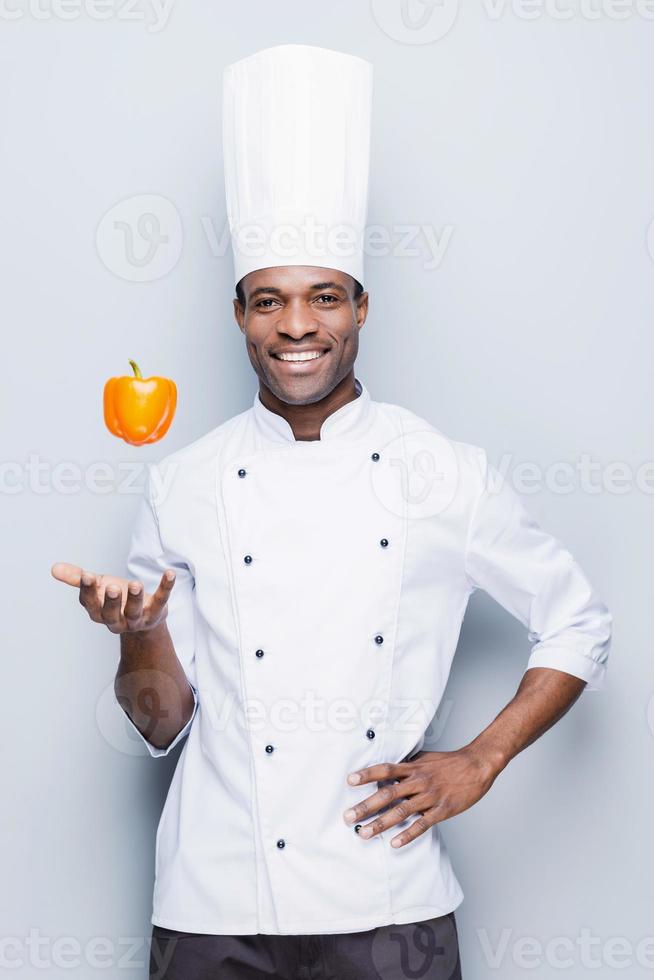 verspielter Kochmeister. selbstbewusster junger afrikanischer koch in weißer uniform, der pfeffer wirft und lächelnd in die kamera blickt, während er vor grauem hintergrund steht foto