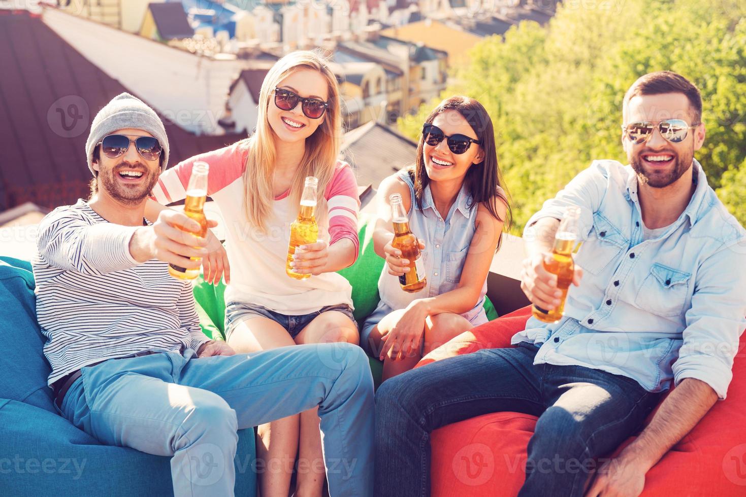 Prost Draufsicht von vier jungen, fröhlichen Menschen, die Flaschen mit Bier halten und mit Ihnen jubeln, während sie an den Sitzsäcken auf dem Dach des Gebäudes sitzen foto