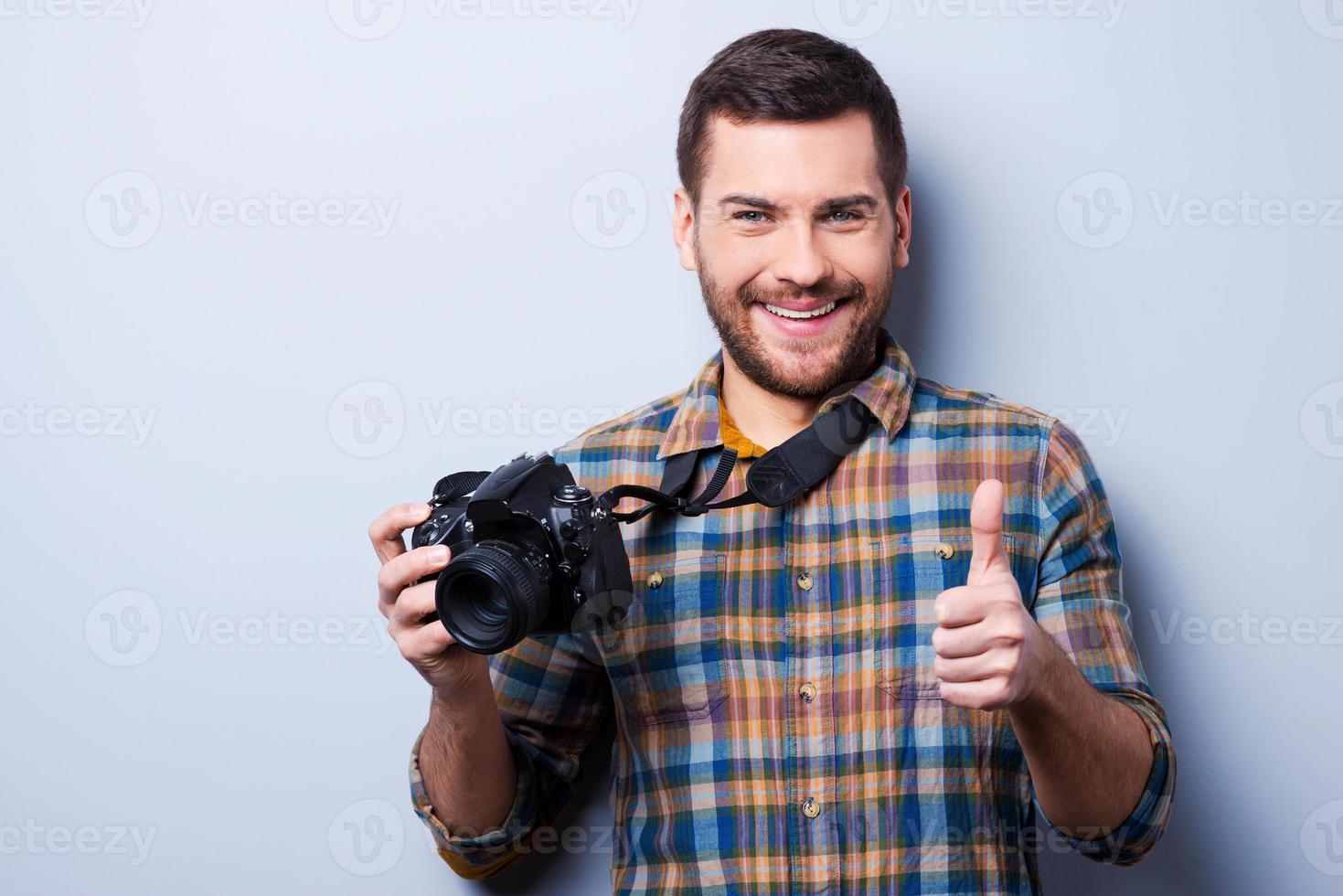 Gutes Lächeln Porträt eines selbstbewussten jungen Mannes im Hemd, der die Kamera hält und den Daumen nach oben zeigt, während er vor grauem Hintergrund steht foto