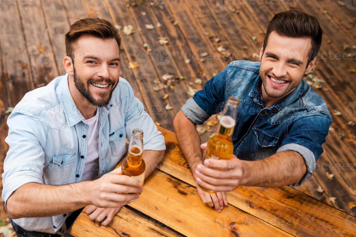 Prost Draufsicht von zwei fröhlichen jungen Männern, die Flaschen mit Bier ausstrecken und die Kamera betrachten, während sie im Freien stehen foto