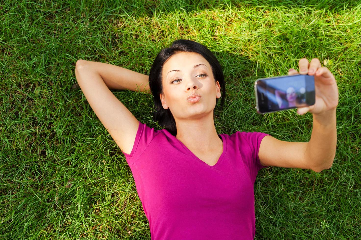 frau, die selfie macht. Blick von oben auf die schöne junge Frau, die mit ihrem Smartphone ein Selfie macht, während sie im grünen Gras liegt foto