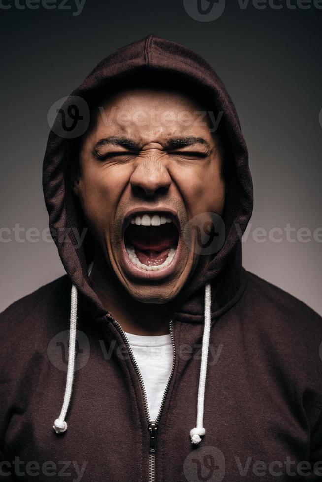 Energie in ihm. Wütender junger afrikanischer Mann im Kapuzenhemd, der die Augen geschlossen hält und schreit, während er vor grauem Hintergrund steht foto