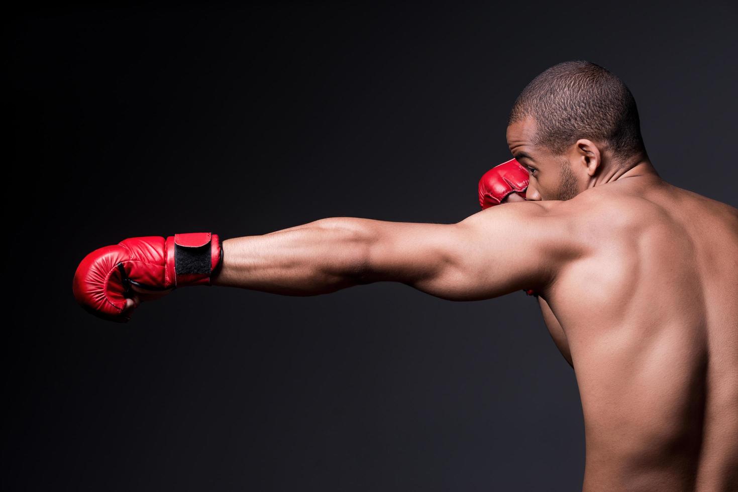 seine Boxkünste trainieren. Seitenansicht eines jungen, hemdlosen Afrikaners in Boxhandschuhen, der im Stehen vor grauem Hintergrund trainiert foto