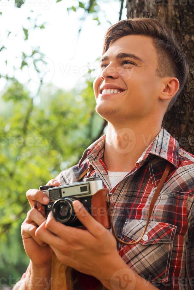 glücklicher Fotograf. hübscher junger Mann, der eine Vintage-Kamera hält und lächelt, während er sich an den Baum lehnt foto