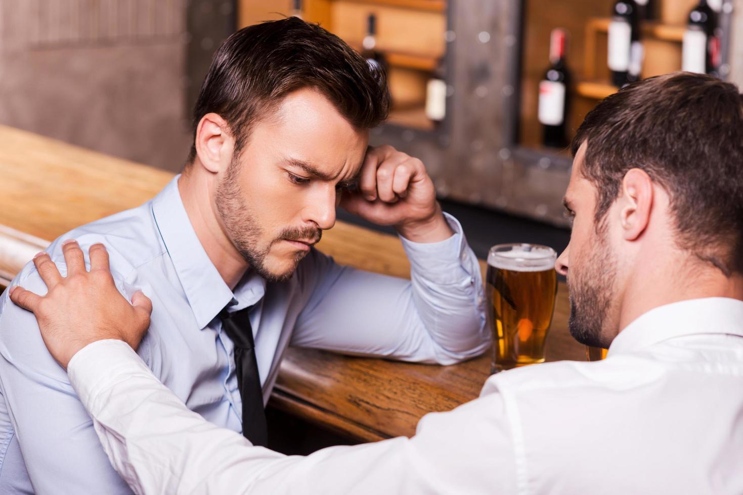 tröstender Freund. frustrierter junger mann in hemd und krawatte, der von seinem freund in der bar getröstet wird foto