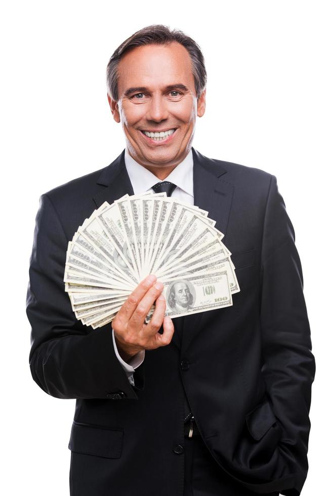 wohlhabend und erfolgreich. Oberkörperaufnahme eines selbstbewussten, reifen Mannes in Abendkleidung, der Geld hält und lächelt, während er vor weißem Hintergrund steht foto