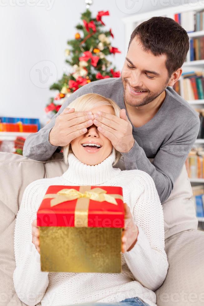 Überraschen Sie die schöne junge Frau, die auf der Couch sitzt und eine Geschenkbox hält, während ihr Freund hinter ihr steht und ihre Augen mit den Händen bedeckt foto