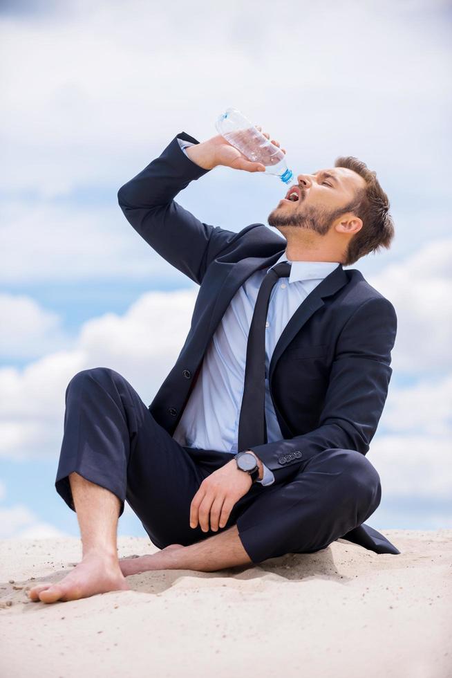 erschöpft und durstig. Junger Geschäftsmann trinkt Wasser, während er auf der Spitze der Sanddüne sitzt foto