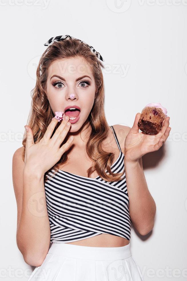 Gesprengte schöne junge Frau, die einen kleinen Kuchen hält und ihren Mund mit der Hand bedeckt, während sie vor weißem Hintergrund steht foto