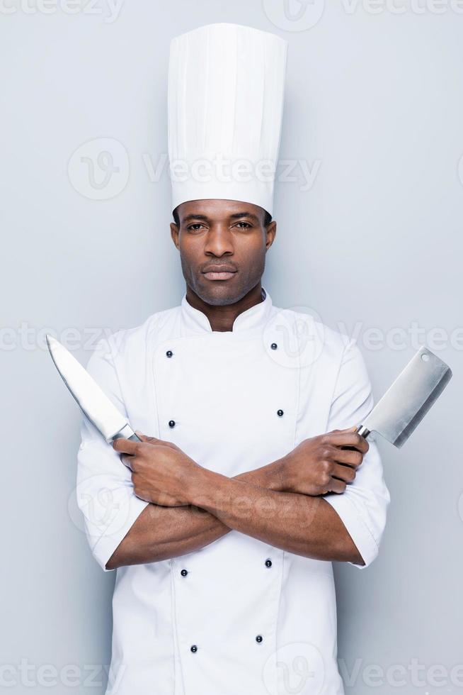 bereit zu arbeiten. selbstbewusster junger afrikanischer koch in weißer uniform, der messer in den händen hält und in die kamera schaut, während er vor grauem hintergrund steht foto