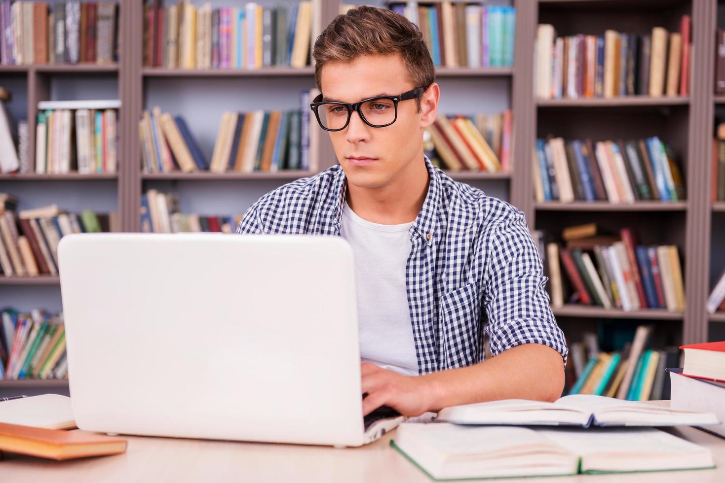 Prüfungsvorbereitung in der Bibliothek. Selbstbewusster junger Mann, der am Laptop arbeitet, während er am Schreibtisch und vor dem Bücherregal sitzt foto