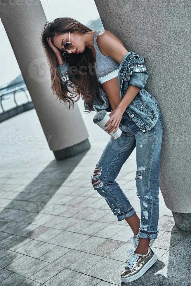 unbeschwerte Schönheit. volle Länge der attraktiven jungen Frau in der Jeansabnutzung, die einen Einwegbecher hält und die Hand im Haar hält, während sie sich draußen auf die architektonische Säule lehnt foto