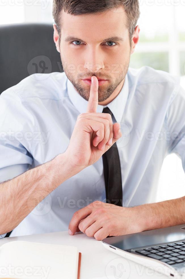 schweigen Sie selbstbewusster junger Mann in Abendkleidung, der den Finger auf den Mund hält und in die Kamera schaut, während er an seinem Arbeitsplatz sitzt foto