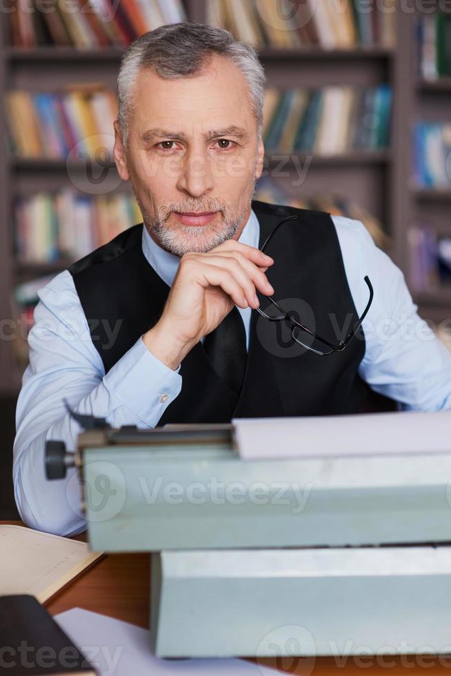 nur inspiriert. Selbstbewusster älterer Mann mit grauem Haar in Abendkleidung, der an der Schreibmaschine sitzt und mit einem Bücherregal im Hintergrund in die Kamera blickt foto