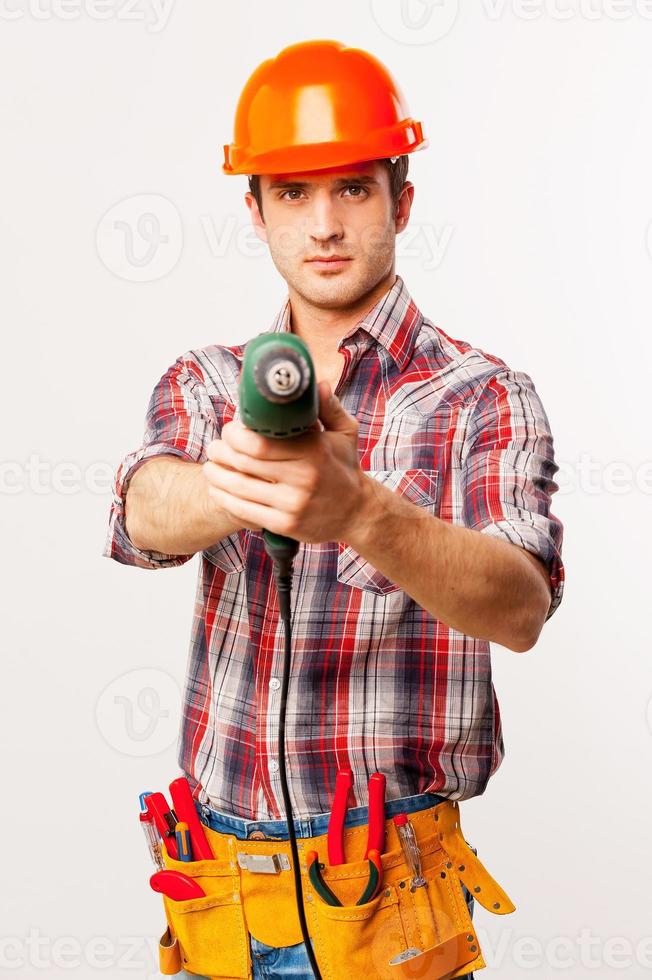 Frieren Sie den hübschen jungen Heimwerker im Helm aus, der Bohrer ausdehnt und die Kamera anschaut, während er vor grauem Hintergrund steht foto