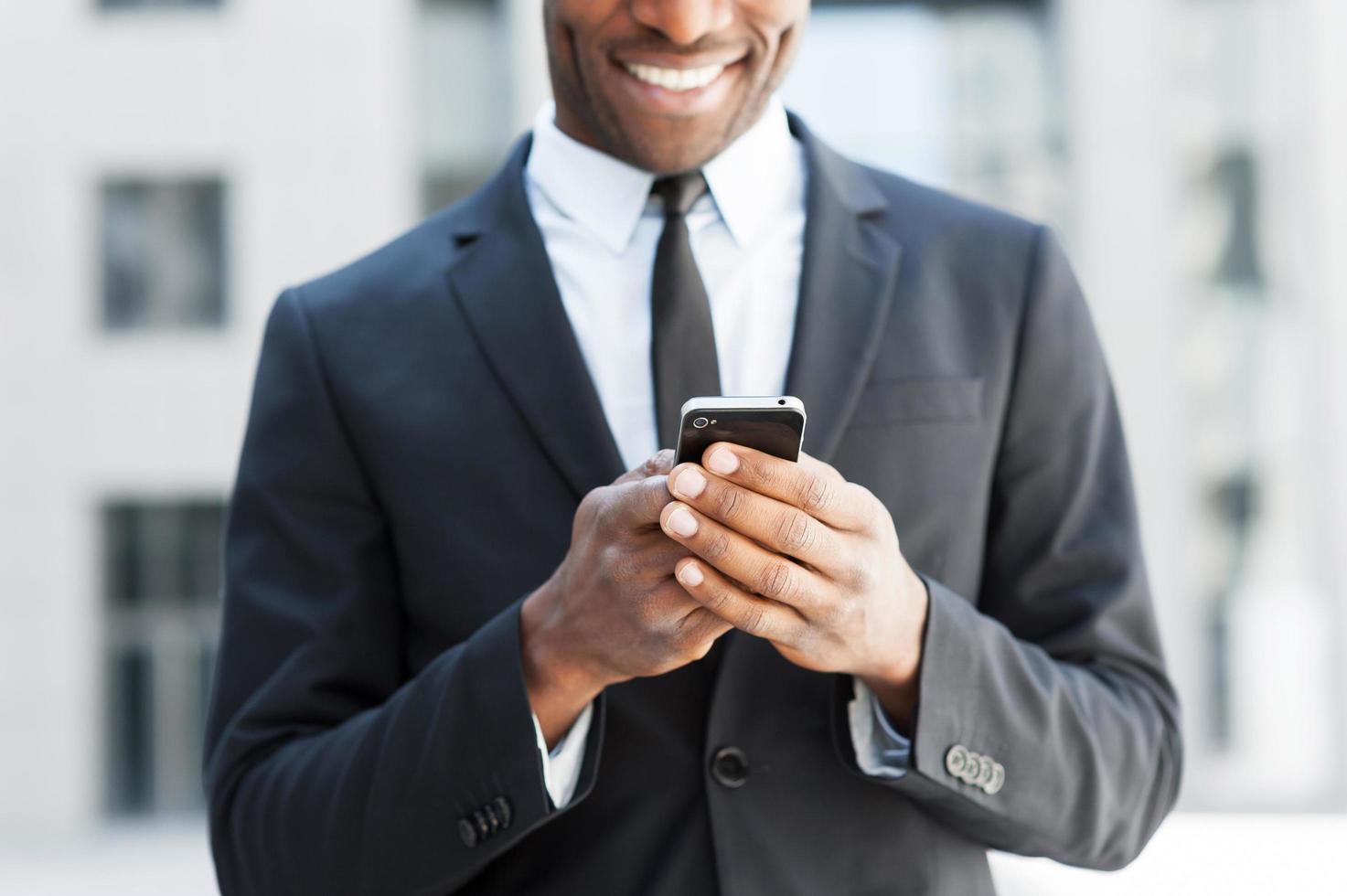 er ist immer in Kontakt. Zugeschnittenes Bild eines fröhlichen jungen afrikanischen Mannes in Abendkleidung, der ein Handy hält und lächelt, während er im Freien steht foto