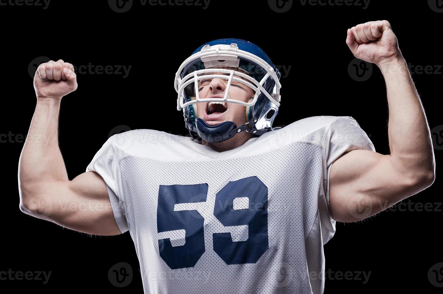 Sieg amerikanischer Fußballspieler schreit und hält die Arme erhoben, während er vor schwarzem Hintergrund steht foto