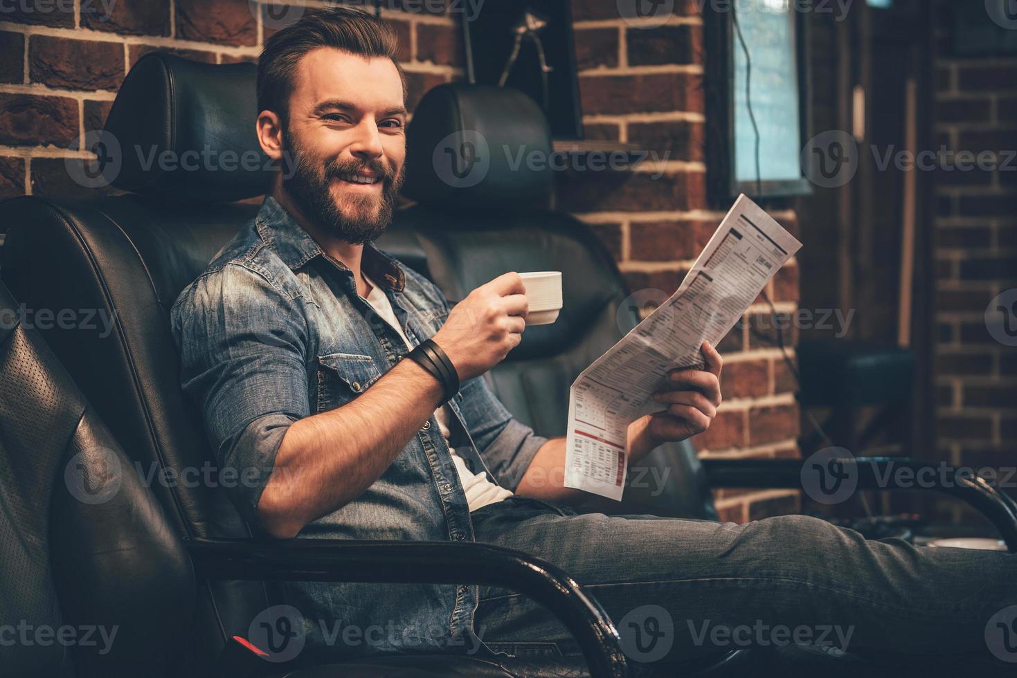 jetzt ist dies ein gutaussehender junger bärtiger mann, der mit einem lächeln in die kamera schaut und eine tasse kaffee und zeitung hält, während er in einem bequemen stuhl im friseursalon sitzt foto