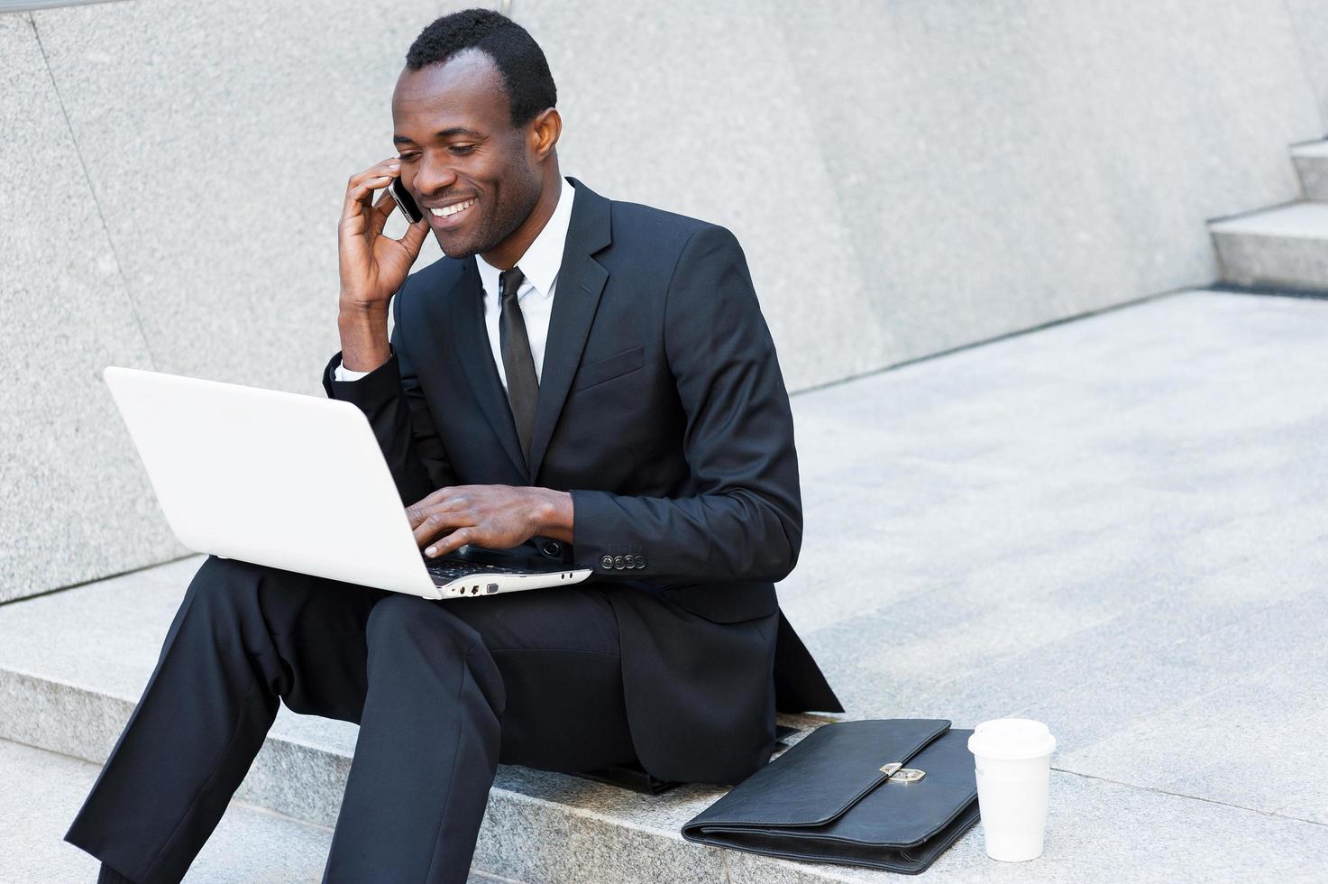 Geschäft ist sein Leben. Fröhlicher junger afrikanischer Mann in Abendkleidung, der am Handy spricht und am Laptop arbeitet, während er auf der Treppe im Freien sitzt foto