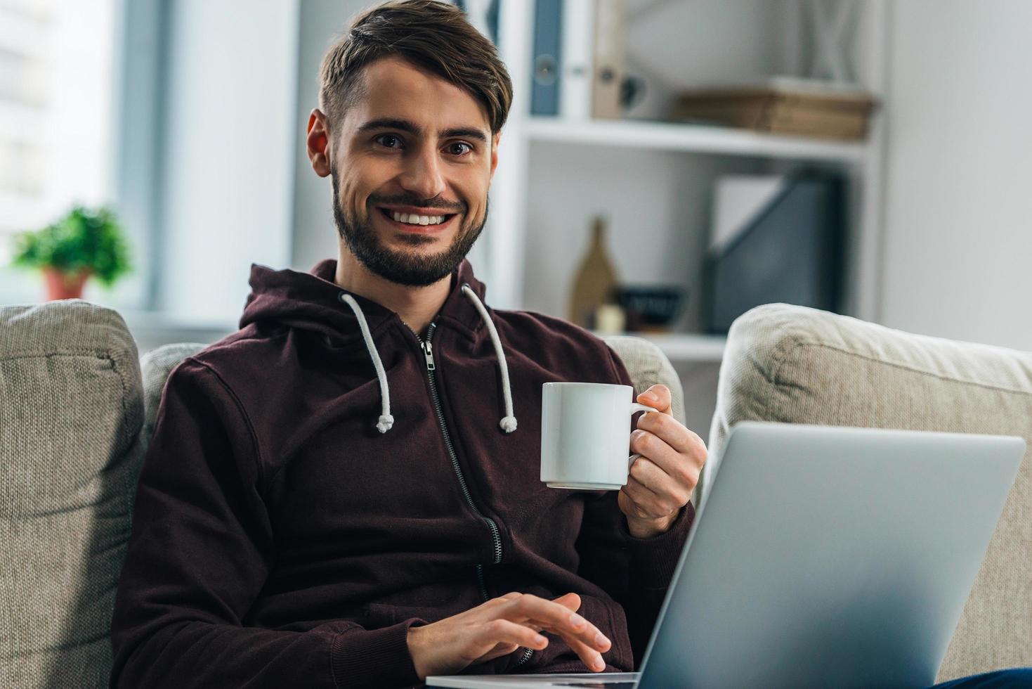 Surfen im Netz zu Hause. Fröhlicher junger Mann, der seinen Laptop benutzt und mit einem Lächeln in die Kamera schaut, während er zu Hause auf der Couch sitzt foto