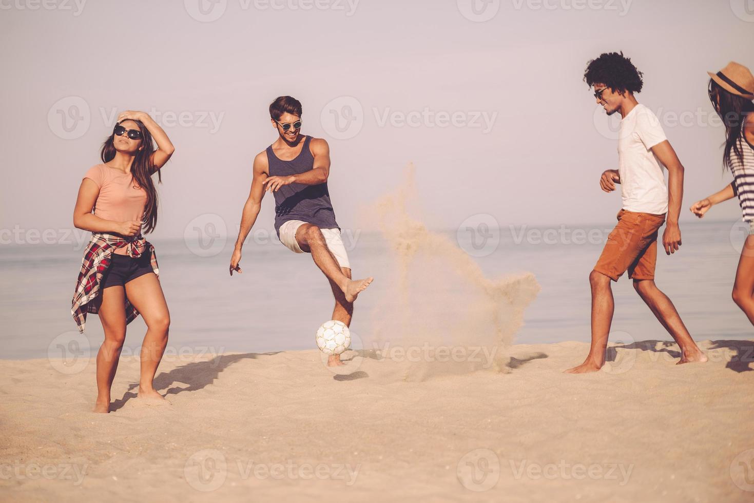 sommerspaß mit freunden. gruppe fröhlicher junger leute, die mit fußball am strand mit meer im hintergrund spielen foto
