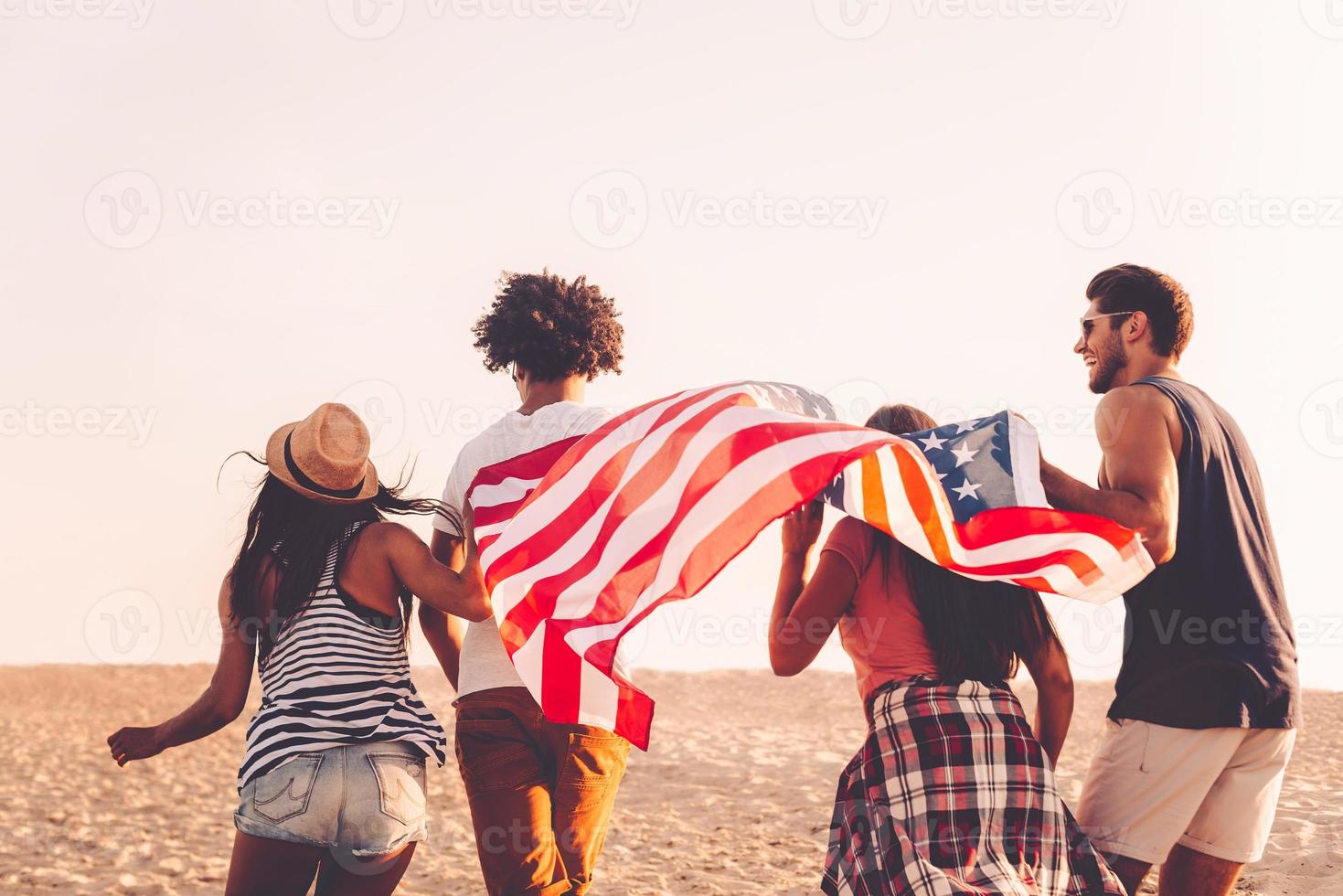 Jung und frei. Rückansicht von vier jungen Menschen, die beim Laufen im Freien die amerikanische Flagge tragen foto