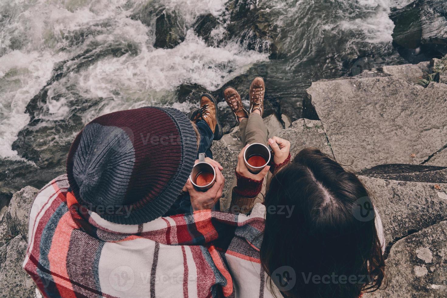 gemeinsam aufwärmen. Nahaufnahme von oben auf ein junges Paar, das mit einer Decke bedeckt ist, die Tassen hält, während es auf den Felsen in der Nähe des Flusses sitzt foto