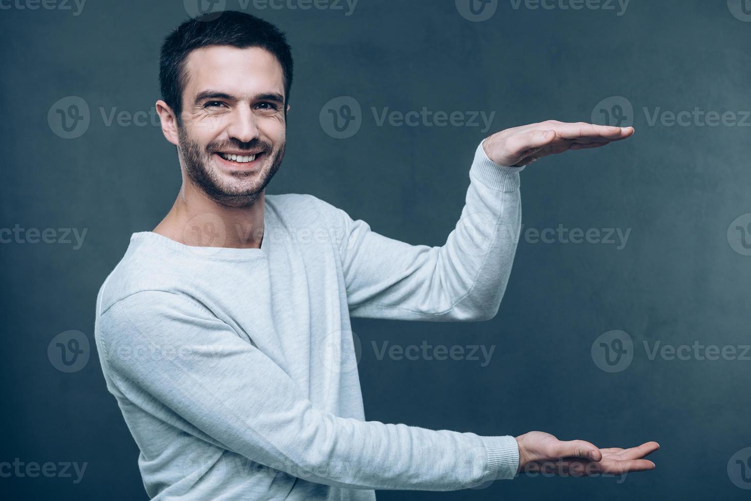 Platz in seinen Händen kopieren. hübscher junger Mann, der Kopienraum hält und die Kamera mit einem Lächeln betrachtet, während er vor grauem Hintergrund steht foto