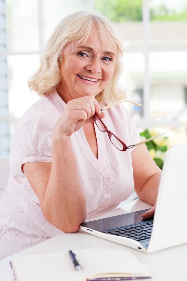 sich jung und energisch fühlen. glückliche Seniorin, die einen Laptop benutzt und in die Kamera lächelt, während sie am Tisch sitzt foto