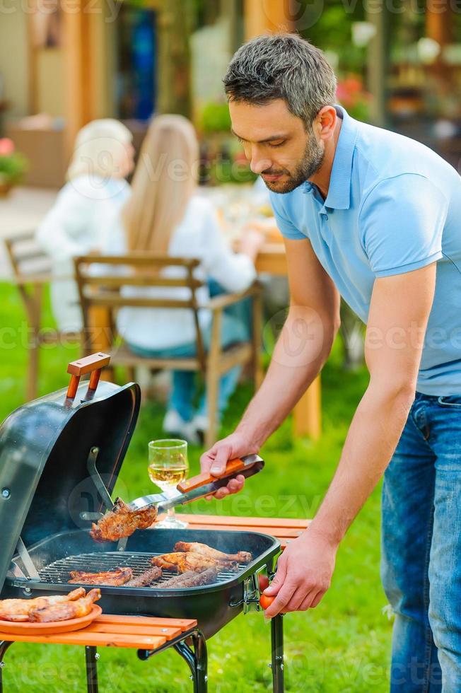 Fleisch perfekt grillen. Selbstbewusster junger Mann, der Fleisch auf dem Grill grillt, während andere Familienmitglieder im Hintergrund am Esstisch sitzen foto