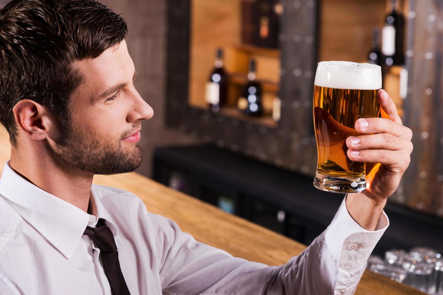 das beste Bier aller Zeiten genießen. Seitenansicht eines hübschen jungen Mannes in Hemd und Krawatte, der Glas mit Bier untersucht und lächelt, während er an der Bartheke sitzt foto