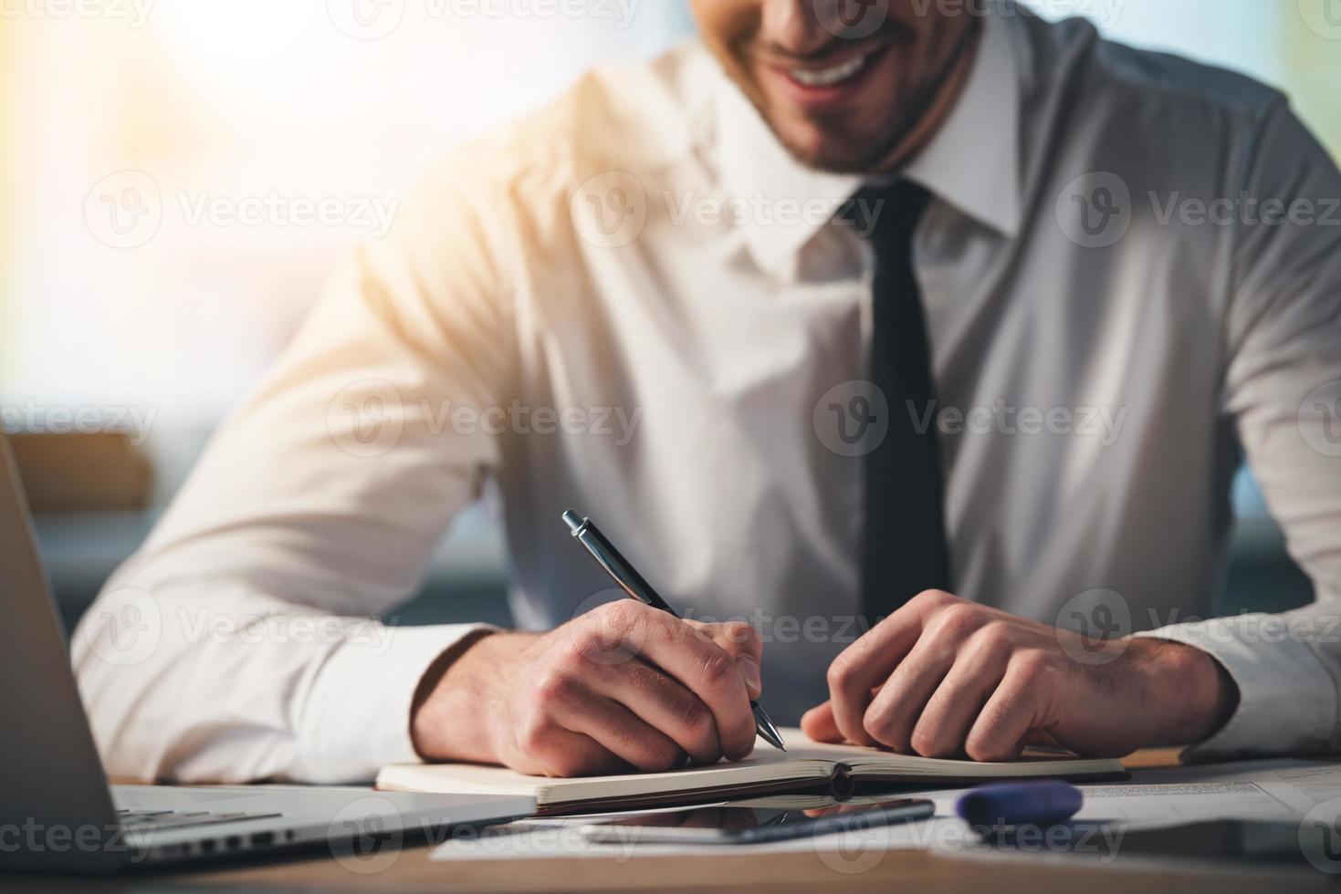 mit Freude arbeiten. Nahaufnahme eines jungen Mannes, der mit einem Lächeln in sein Notizbuch schreibt, während er an seinem Arbeitsplatz sitzt foto