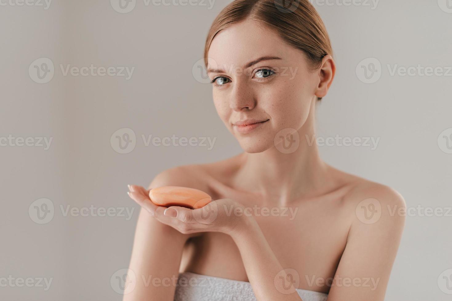 perfekte Körperpflege. Schöne junge Frau mit Sommersprossen im Gesicht, die Seife hält und in die Kamera schaut, während sie vor dem Hintergrund steht foto