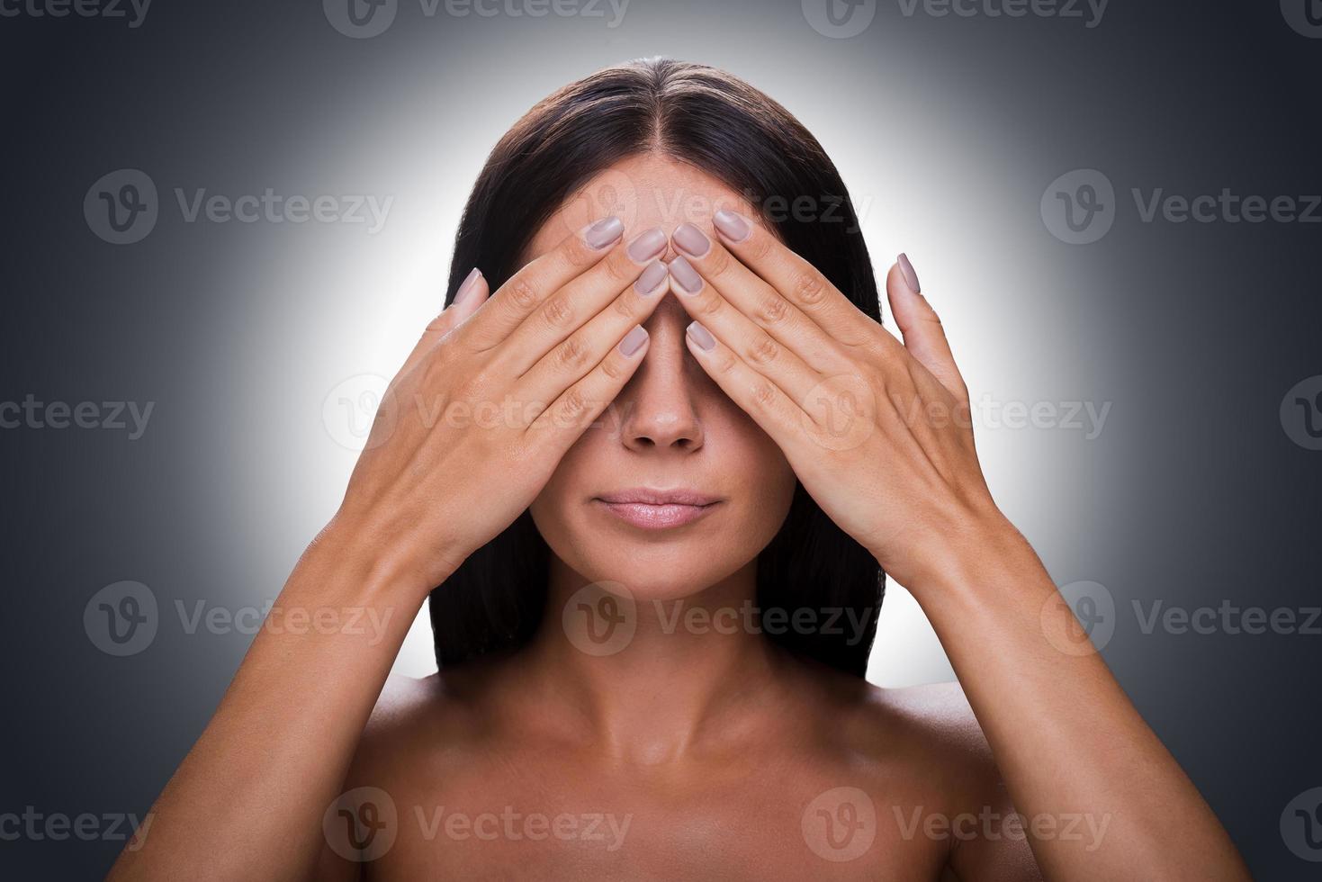 nichts sehen Porträt einer jungen, hemdlosen Frau, die die Augen mit den Händen bedeckt, während sie vor grauem Hintergrund steht foto