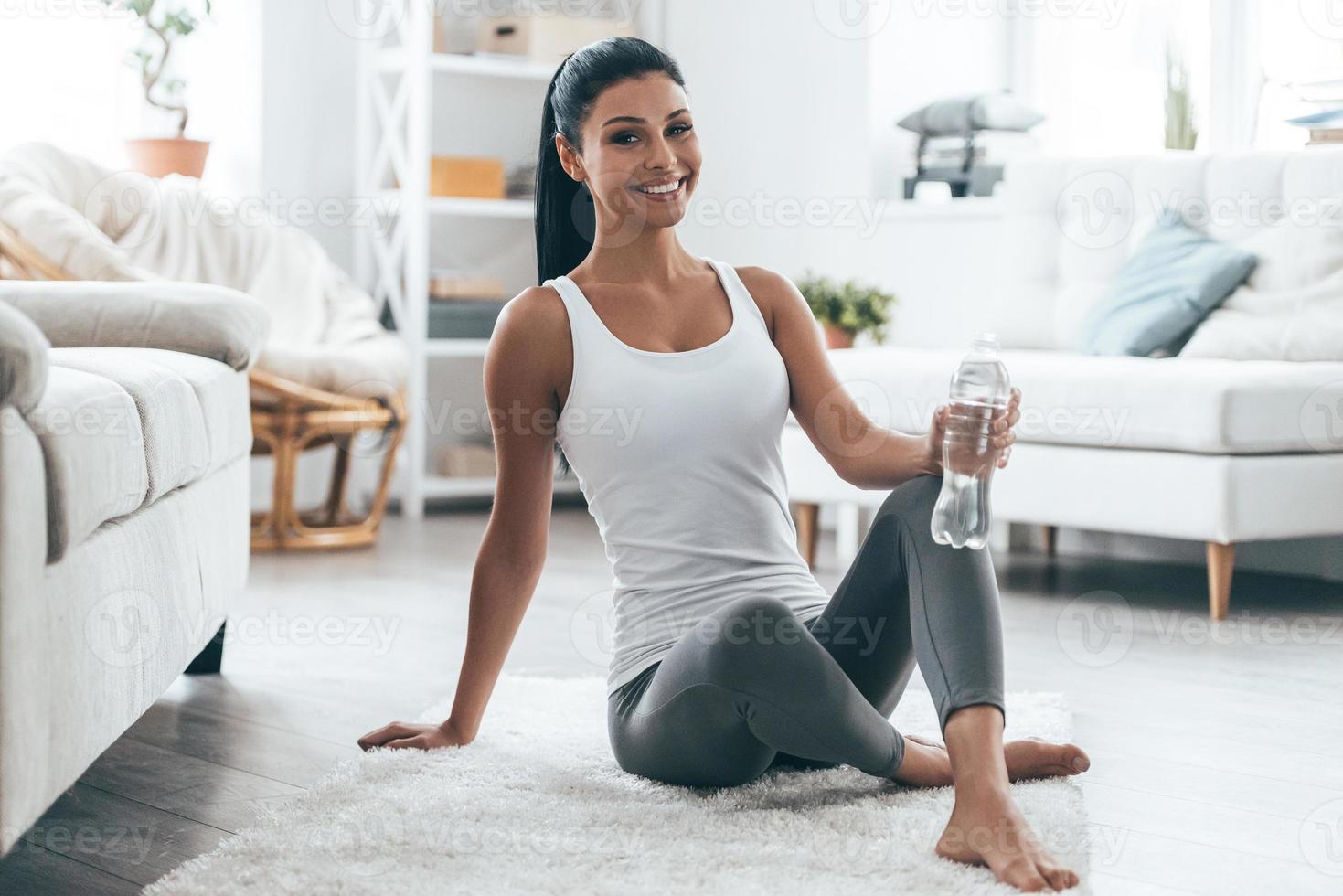 gute arbeit attraktive junge frau, die lächelt und in die kamera schaut, während sie zu hause auf dem teppich sitzt foto