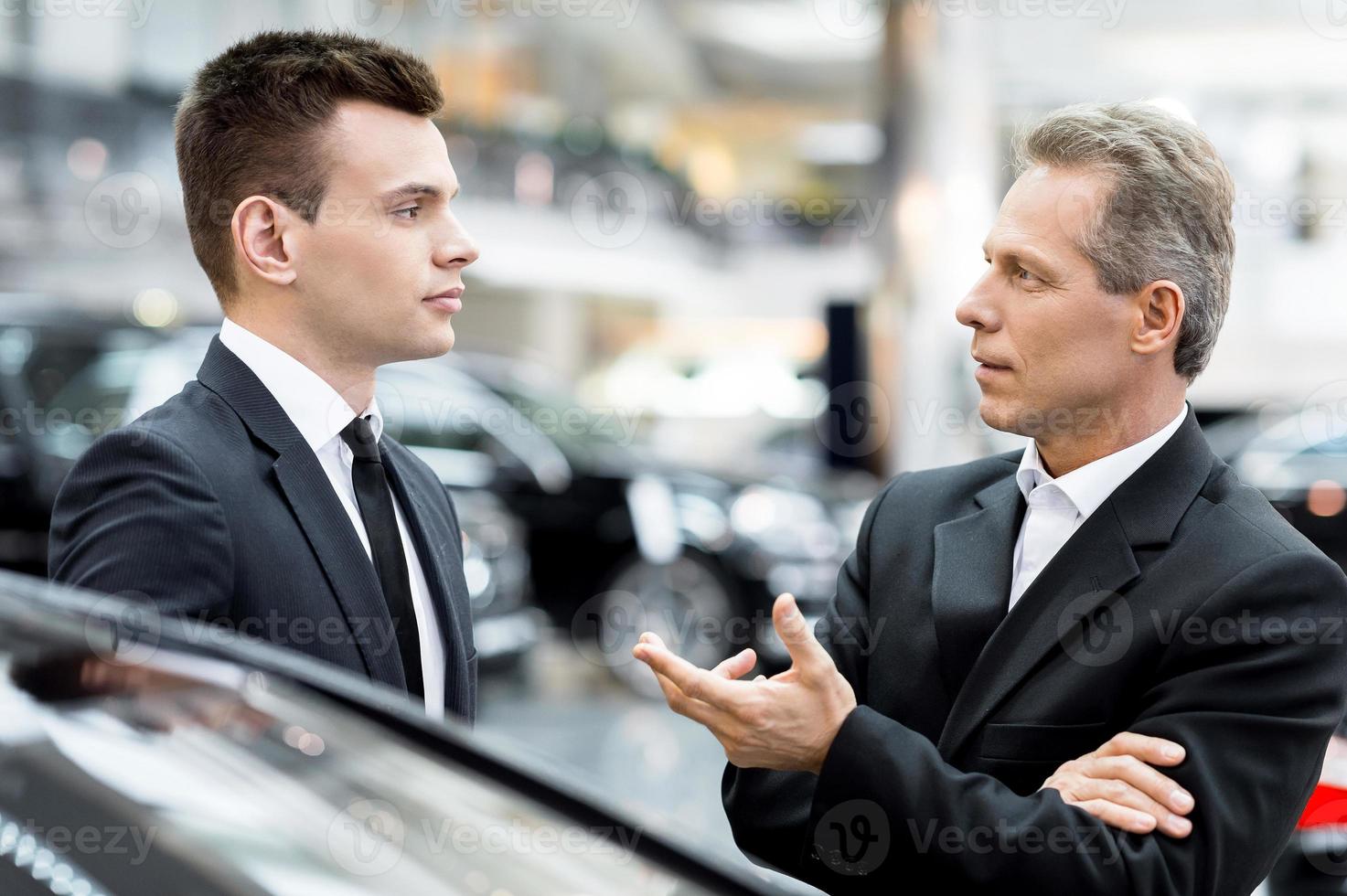 Fahrzeugeigenschaften besprechen. zwei Personen in festlicher Kleidung, die sich unterhalten und gestikulieren, während sie am Autohaus stehen foto