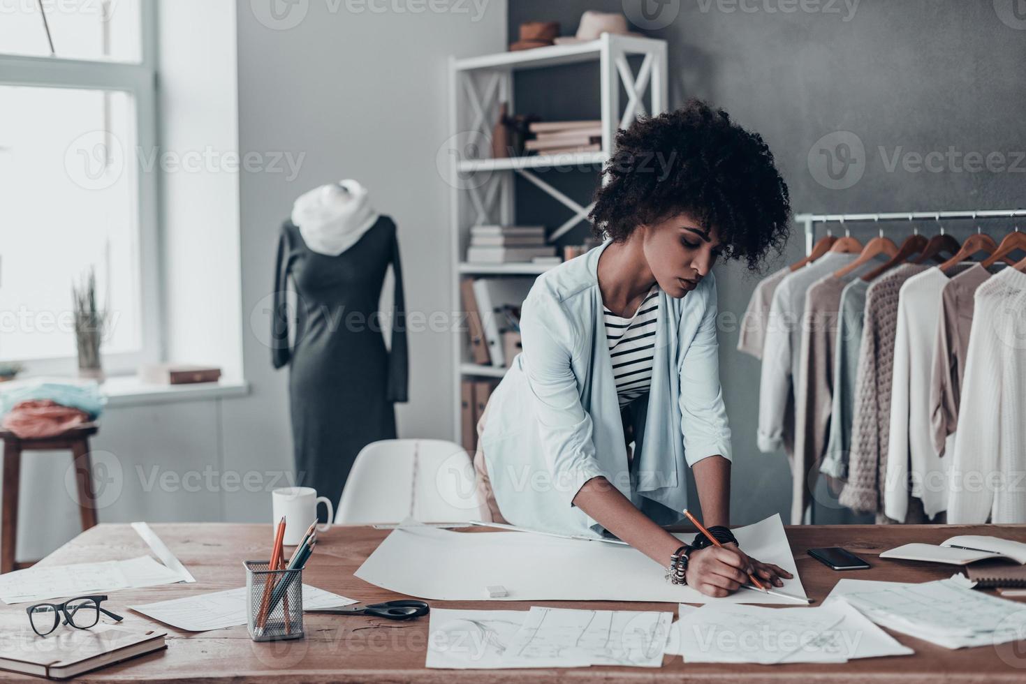 Ideen in Kleidung verwandeln. schöne junge afrikanerin, die an skizzen arbeitet, während sie in ihrem studio in der nähe der kleidung steht, die an den regalen hängt foto