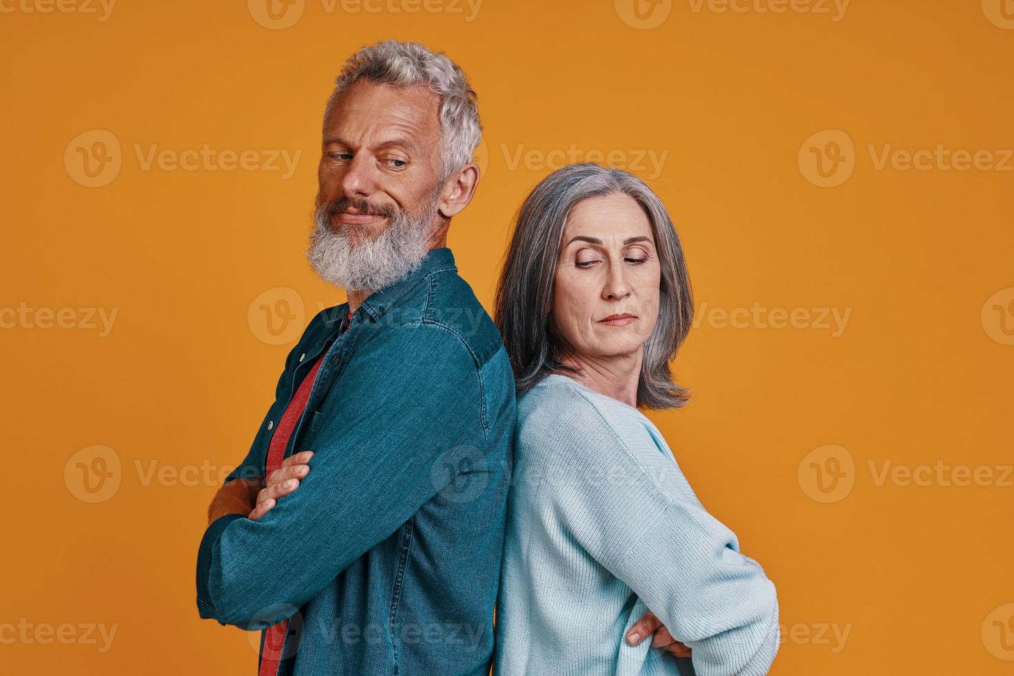 Unzufriedenes älteres Paar, das die Arme verschränkt hält, während es zusammen vor orangefarbenem Hintergrund steht foto