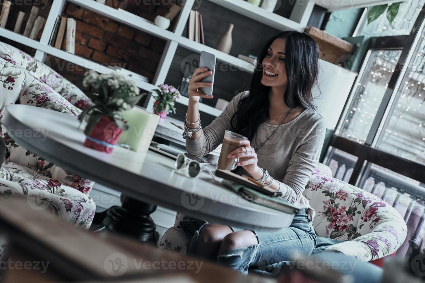 nette Nachrichten bekommen. attraktive junge lächelnde frau, die ihr smartphone benutzt, während sie im restaurant sitzt foto