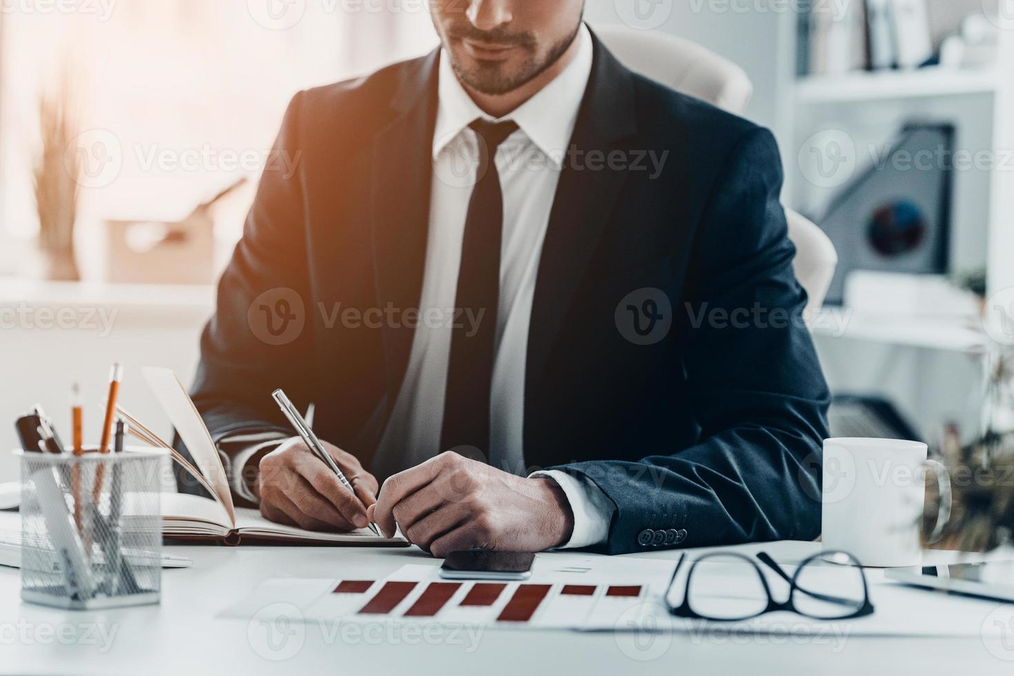schnelle Notizen. Nahaufnahme eines jungen Mannes im vollen Anzug, der etwas in sein Notizbuch schreibt, während er am Schreibtisch sitzt foto