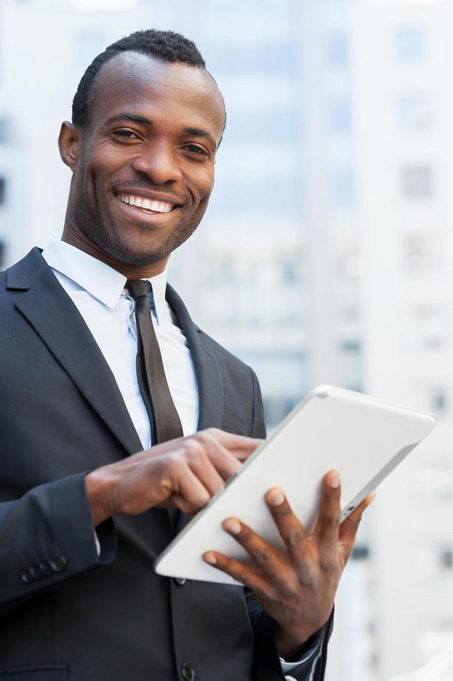 Geschäftsmann aus dem digitalen Zeitalter. Fröhlicher junger afrikanischer Mann in Abendkleidung, der an einem digitalen Tablet arbeitet und lächelt, während er im Freien steht foto
