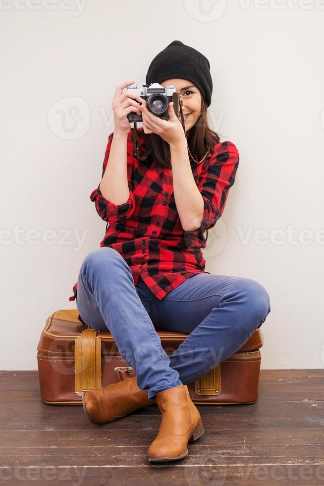 gib mir ein lächeln schöne junge frau in kopfbedeckungen, die eine kamera halten und durch sie schauen, während sie auf einem koffer sitzen foto