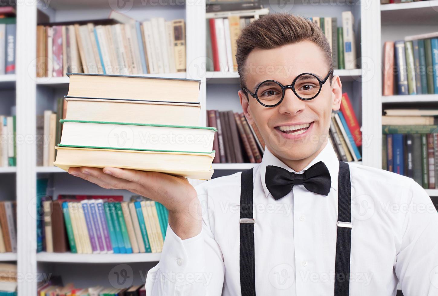 meine Lieblingsbücher. Fröhlicher junger Mann in Hemd und Fliege sitzt am Tisch in der Bibliothek und hält einen Bücherstapel in der Hand foto